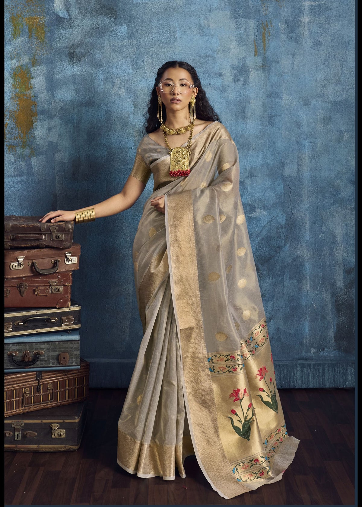 Woman in a saree standing next to stacked suitcases against a textured blue wall.