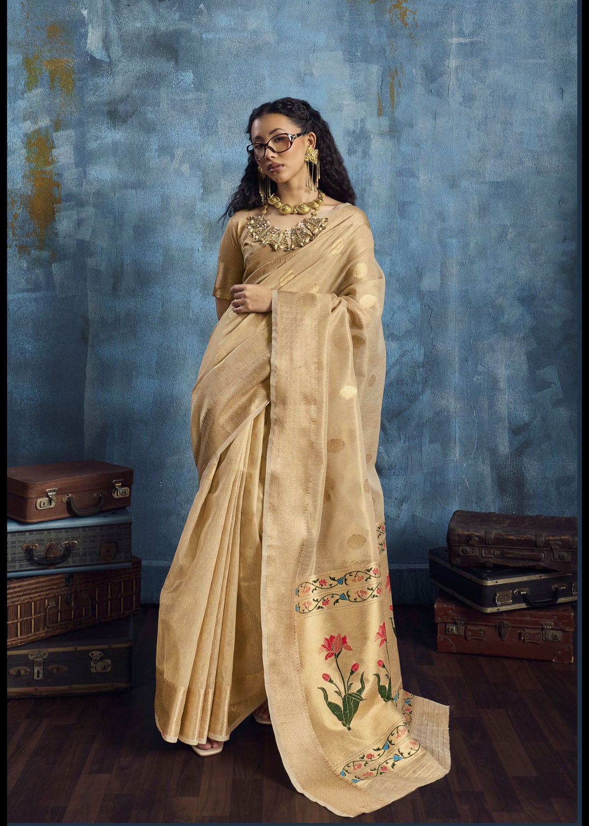 Woman in a beige saree with floral embroidery standing against a textured blue wall.