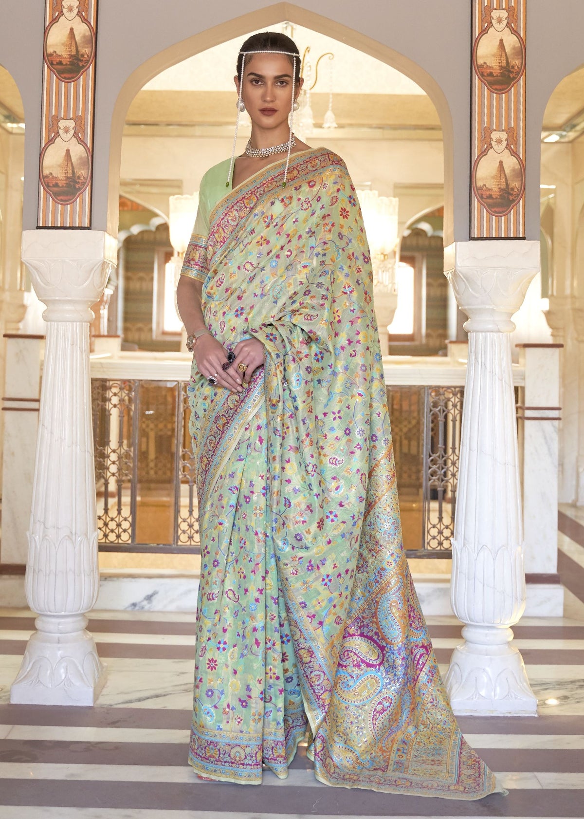 Woman wearing a light green Kashmiri silk saree with multicolored floral and paisley motifs, styled with traditional jewelry in a regal heritage setting.