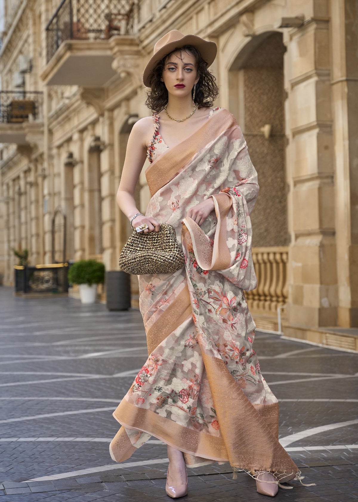 Beige–peach floral saree with modern styling — model walking through city street wearing a soft printed saree with floral straps blouse, paired with a beige hat and pink heels, blending traditional Indian drape with contemporary street fashion.