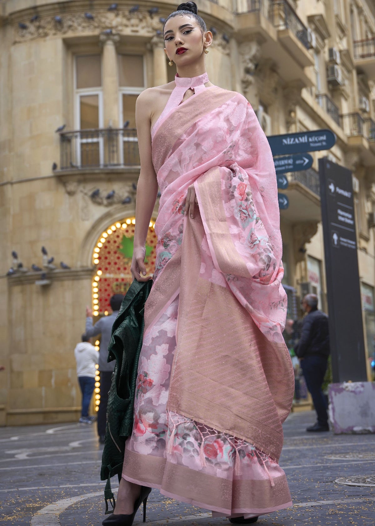 Pink floral saree with halter blouse — model standing on Nizami Street wearing a printed poly brasso saree with intricate floral motifs, paired with black heels and a green drape, styled for bold modern Indian fashion photography.