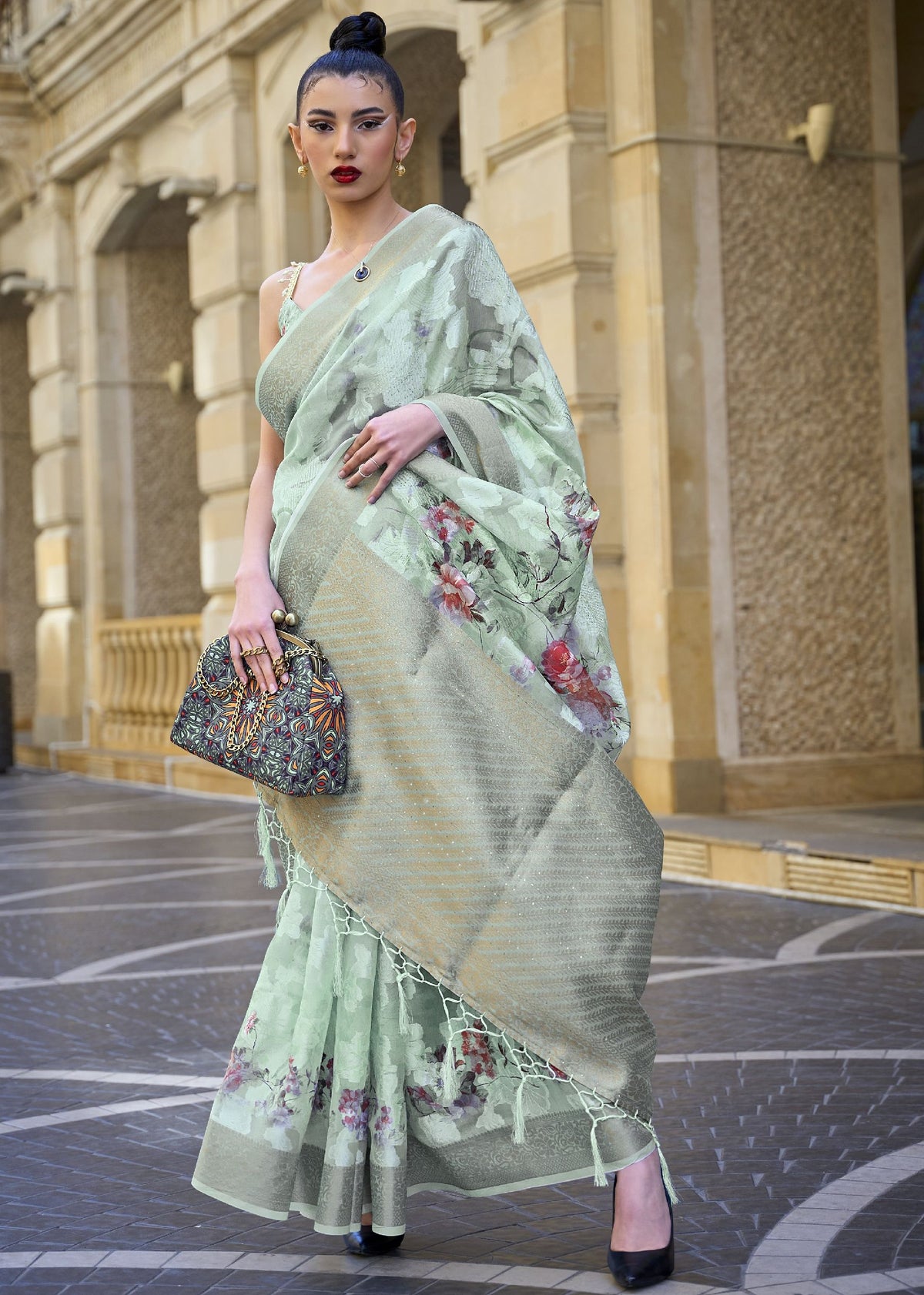 Light green floral saree with gold border — model posing in front of arched architecture wearing a printed poly brasso handloom saree with tassels and embroidered handbag, styled with gold jewellery for a premium ethnic look.