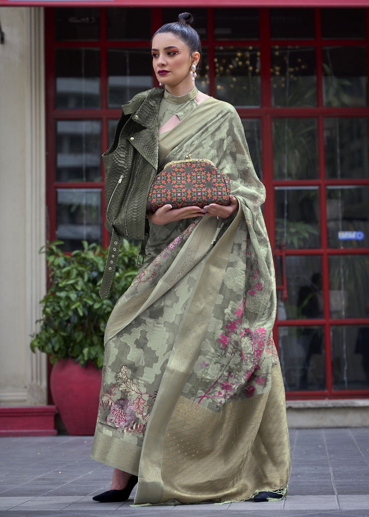 Olive green embroidered saree with jacket — model wearing a floral‑embroidered saree paired with a textured jacket and patterned clutch, photographed in an urban setting for a fusion Indo‑Western fashion aesthetic.
