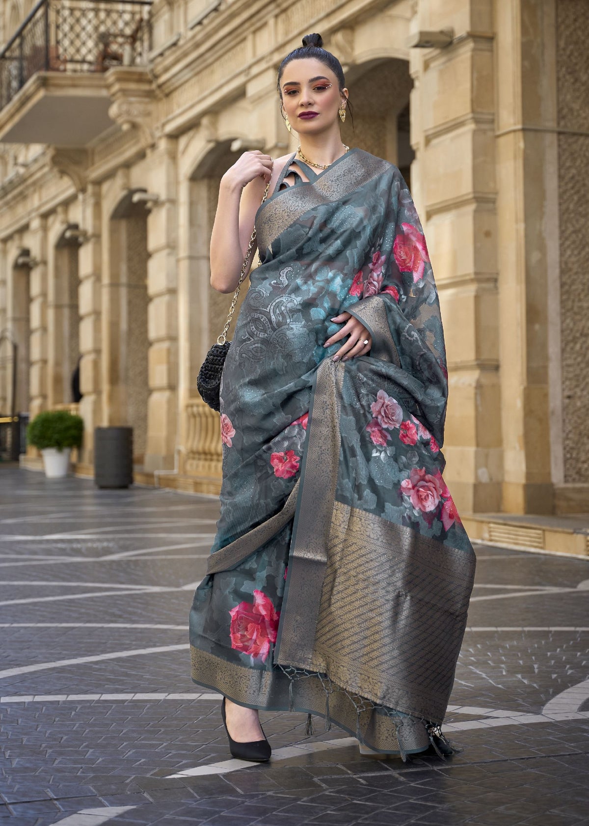Grey–pink floral silk saree with metallic border — model standing in a courtyard wearing a printed poly brasso saree with floral detailing, gold jewellery, and black handbag, styled for luxury Indian fashion photography.