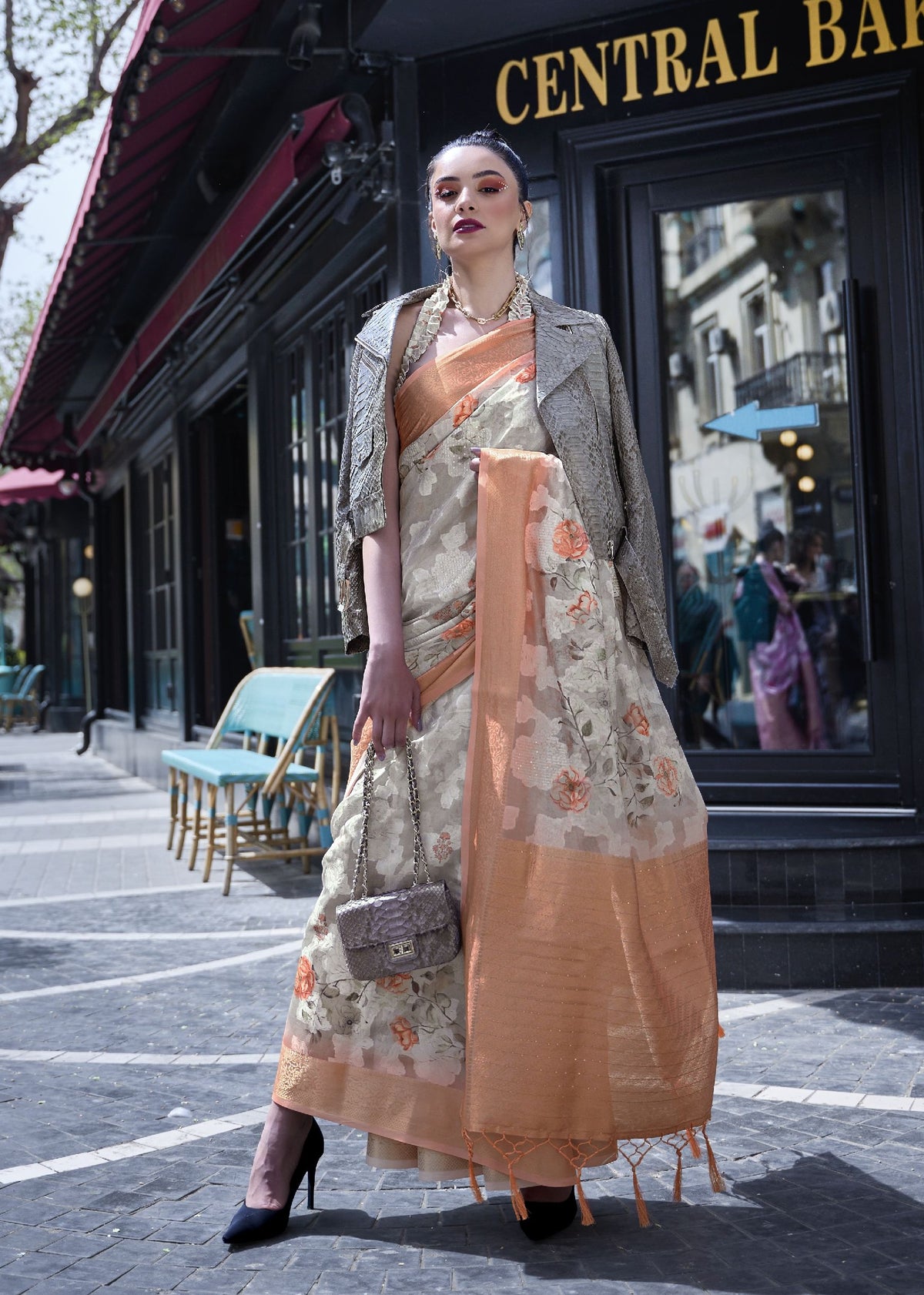 Light blue floral saree with beige pallu — model posing in front of historic balconies wearing a printed poly brasso saree with pink and red floral embroidery, accessorised with statement earrings and a patterned handbag for a cinematic Bollywood look.