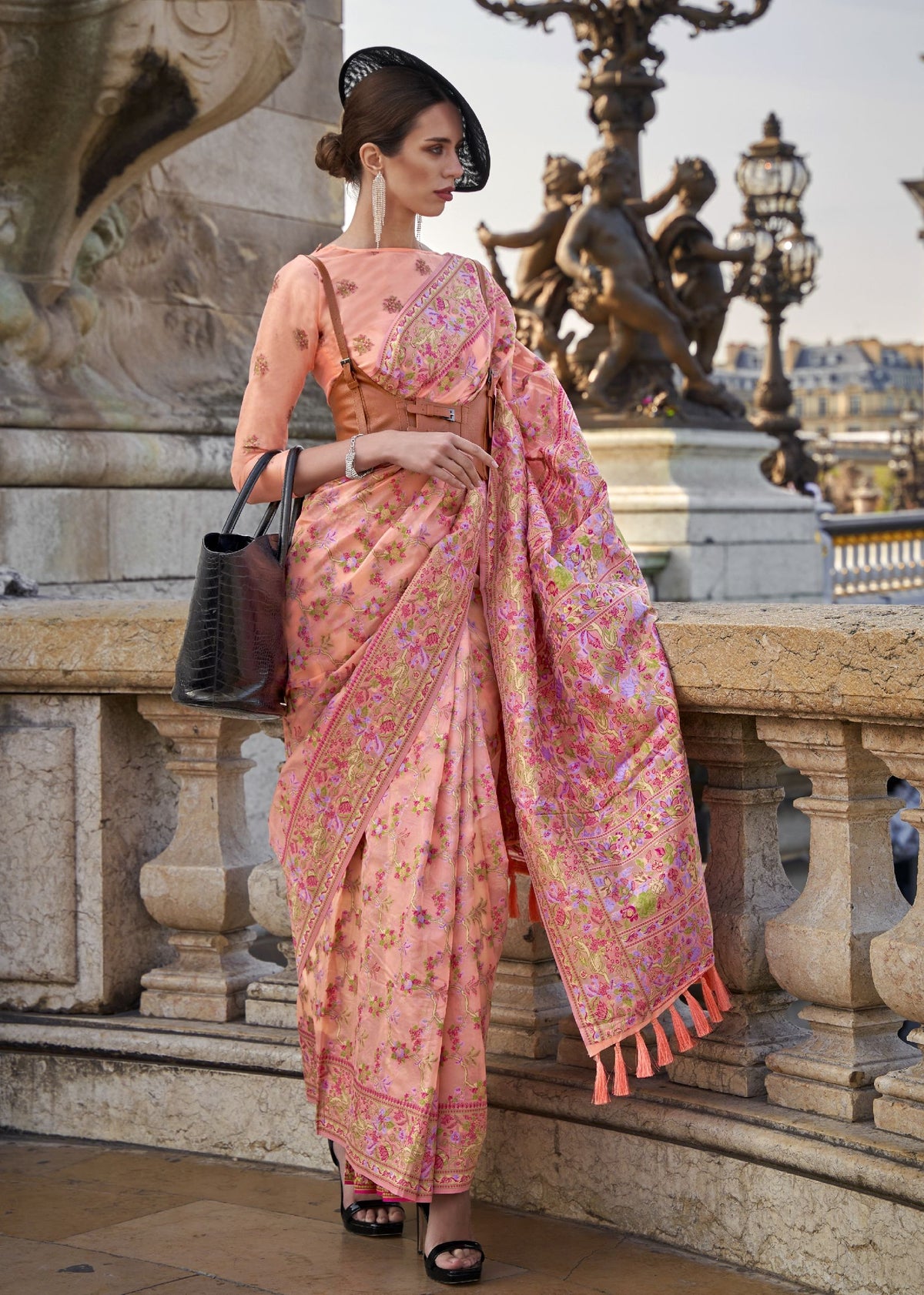 Soft peach saree with detailed pink and green floral embroidery and a gold tasselled border.