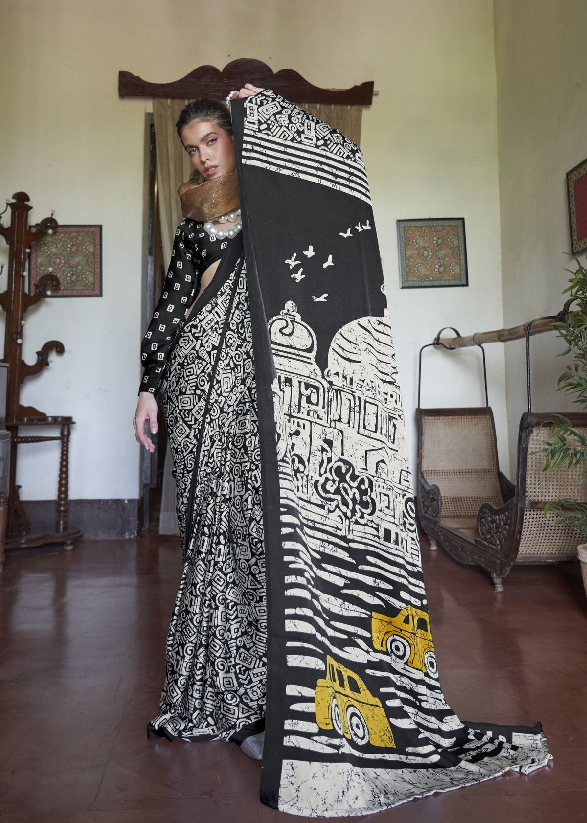 Woman holding a black and white patterned scarf in a room with wooden furniture and decorative , items. Kaalin satin saree - front view